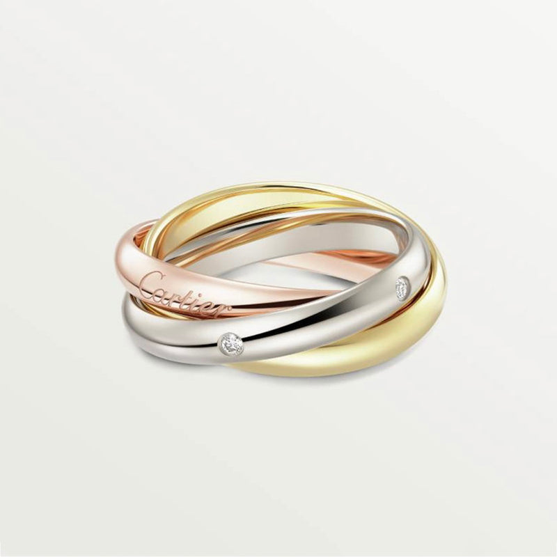 Three interlocking Cartier rings in yellow gold, white gold, and rose gold, with two small clear stones and a visible Cartier logo engraving, on a light gray background.