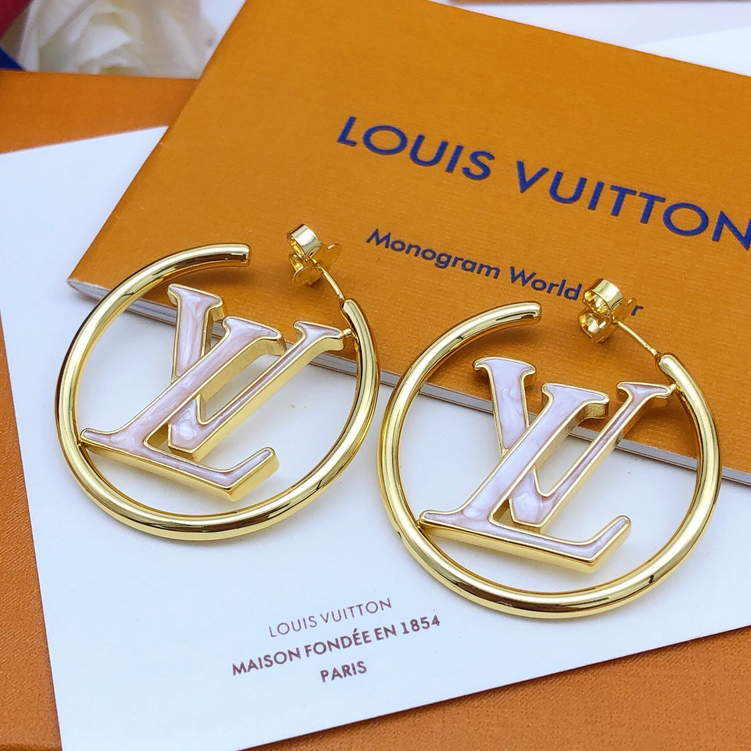 A pair of gold-tone Louis Vuitton hoop earrings with a white enamel monogram LV in the center, displayed on a white card over an orange Louis Vuitton box.