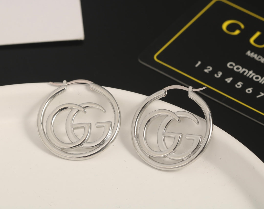 Silver hoop earrings with Gucci logo on a white surface with Gucci packaging in the background.