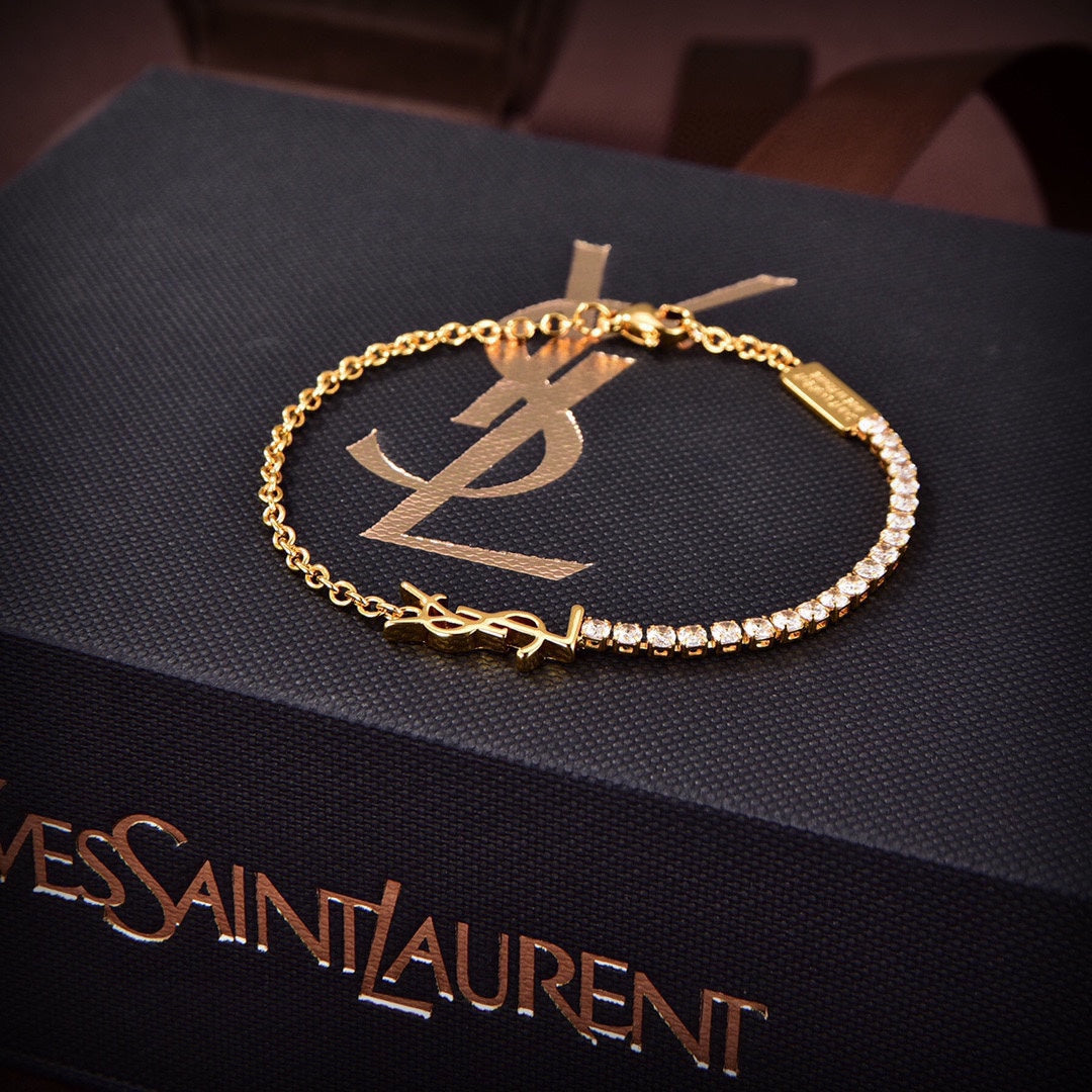 A gold-tone chain bracelet with a YSL logo charm and a row of clear stones, displayed on a black textured Yves Saint Laurent box.